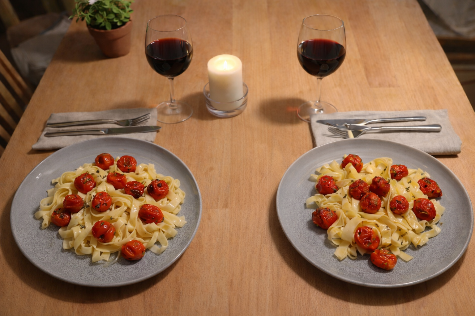 Zwei graue Teller mit Tagliatelle und Cherrytomaten stehen auf einem hellen Holztisch gegenüber voneinander. Daneben liegen Besteck auf beigen Stoffservietten, zwei Rotweingläser und eine brennende Kerze in der Mitte.